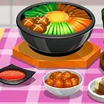Bàn ăn với một nồi lẩu đầy nguyên liệu, cùng với các món phụ như kimchi, đậu phụ, và một ly trà xanh trên khăn trải bàn kẻ ô màu hồng