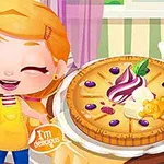 Bé gái tóc vàng mặc tạp dề đứng cạnh chiếc bánh với kem và trái cây trang trí, bên cạnh là điện thoại thông minh