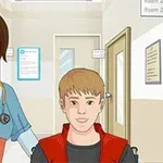 Một y tá cười đứng bên cạnh một cậu bé tóc vàng ngồi trên xe lăn trong hành lang bệnh viện